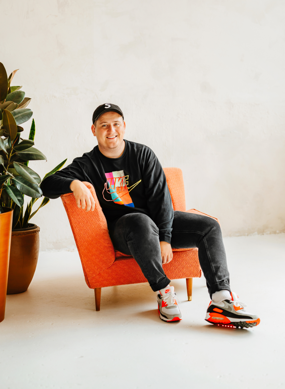 Adam sat on a vintage orange chair smiling wearing a baseball cap, Nike airman sneakers, jeans and a long sleeve Nike T-shirt