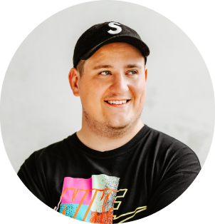 Portrait of Adam smiling with a grey backgorund. Wearing a black baseball cap and a black t-shirt with colourful graphics looking off into the distance.